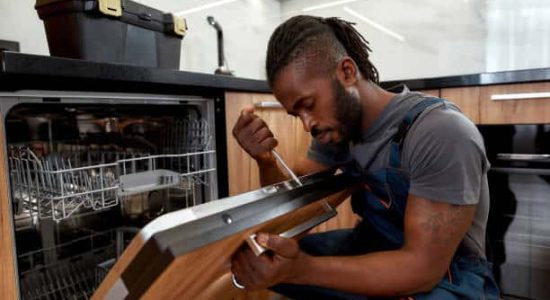 Process of dishwashing machine installation. Young African service man setting up dishwasher in new stylish kitchen. Diverce handyman in workwear, close portrait.